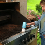 A woman stands next to a stainless steel outdoor grill, using a blue handheld brush to clean the grill grates. The background shows a green garden patio setting.