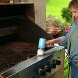 A woman stands next to a stainless steel outdoor grill, using a blue handheld brush to clean the grill grates. The background shows a green garden patio setting.