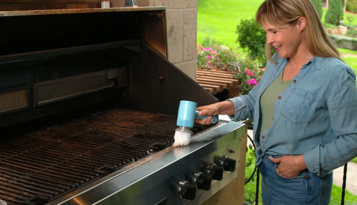 A woman stands next to a stainless steel outdoor grill, using a blue handheld brush to clean the grill grates. The background shows a green garden patio setting.