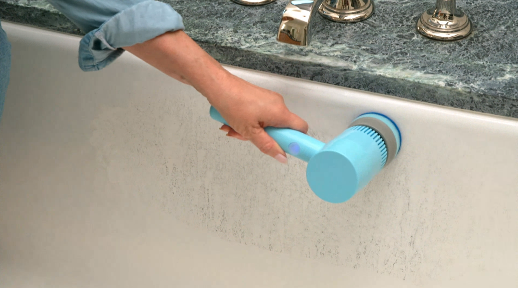 A person using a light blue handheld electric bathtub scrubber on the inside of a white bathtub, with a dark marble countertop and chrome faucet visible in the background.