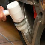 A close-up of a hand using a white electric wheel tire brush to clean the inside of a silver car wheel.